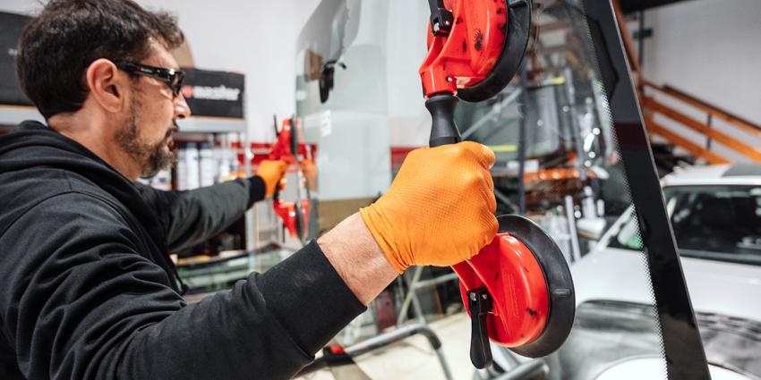 An automotive collision repair technician lifts a new replacement windshield for installation on a vehicle.